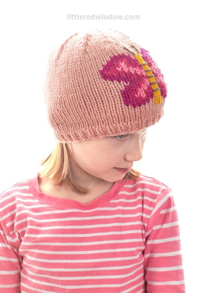 A young child wearing a pink knit hat with a large butterfly design, looking down and to the side. The child is also wearing a pink and white striped shirt.