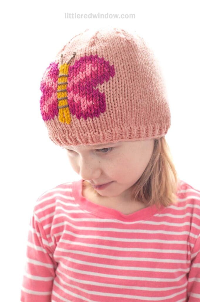 A young child in a pink and white striped shirt wears a light pink knit hat with a large, colorful butterfly design on the front. The child is looking down and smiling slightly.