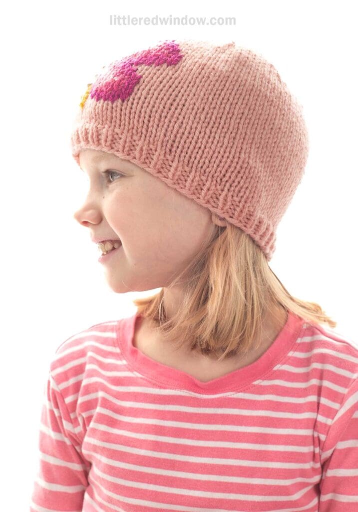 A young girl with blonde hair wearing a pink knit hat with a purple butterfly design and a pink striped shirt, smiling and looking to the left against a white background.