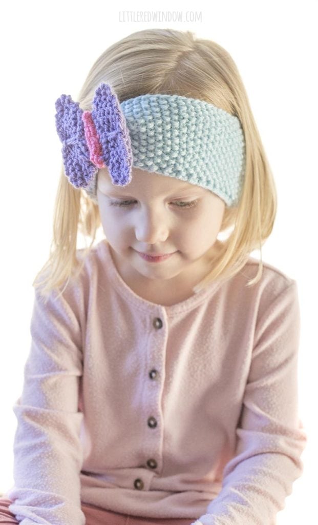 little girl in pink shirt wearing blue knit headband with purple butterfly on the front looking off to the right