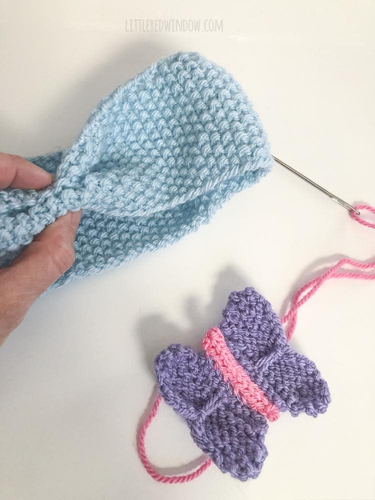 hand pinching a blue knit headband with a knit butterfly on the white table next to it