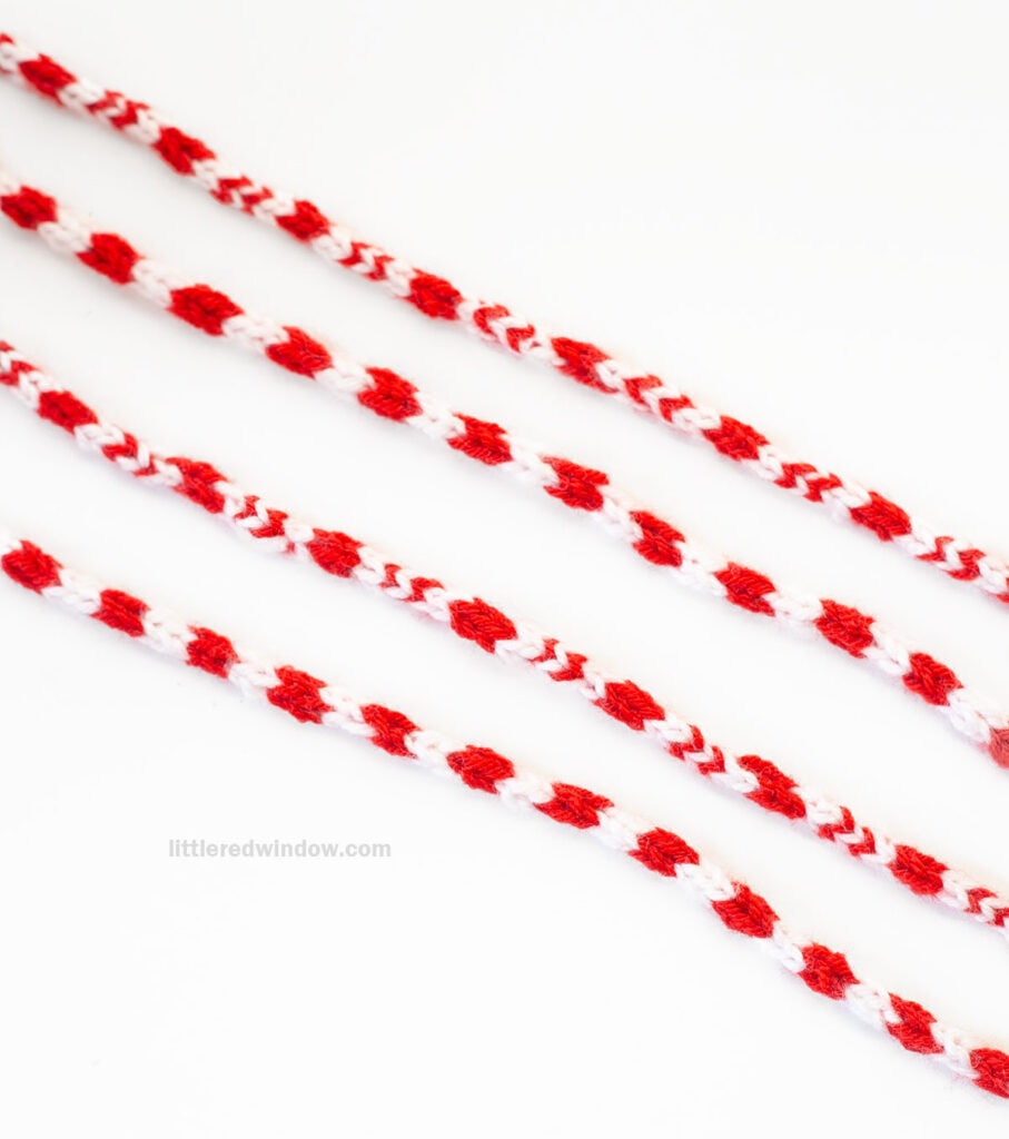 Four strands of red and white candy cane icord are laid out diagonally on a white background. The yarns display a striped pattern with alternating sections of red and white.