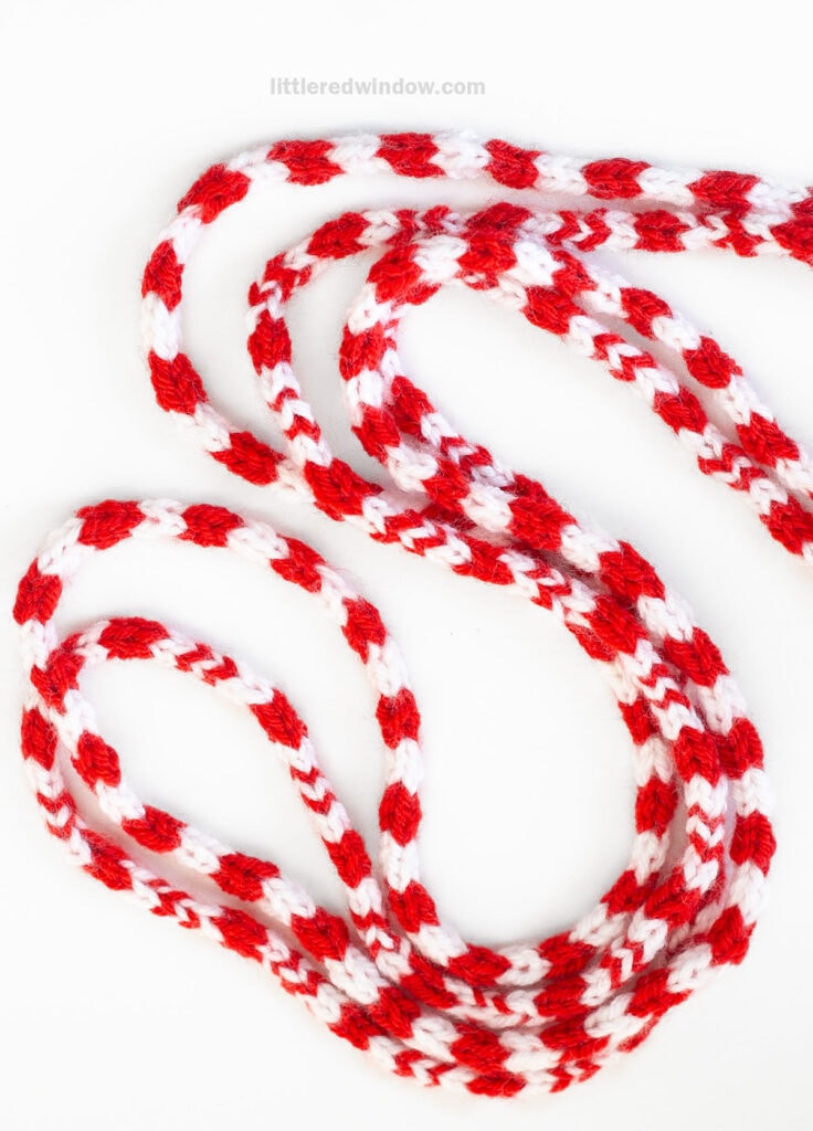 Red and white knitted cord with a striped pattern, resembling a candy cane icord, coiled loosely on a white background.