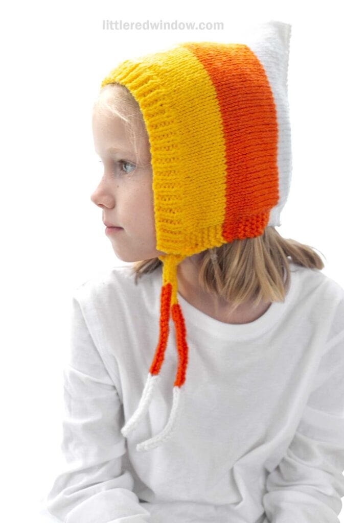 A young child wearing a hand-knitted hat resembling a candy corn, with horizontal stripes in white, orange, and yellow. The child is dressed in a white shirt and is looking to the side with a calm expression.