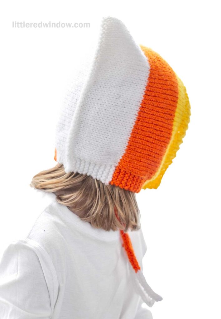 A child wearing a handmade knit hat resembling a candy corn with white, orange, and yellow stripes is shown from the back. The child is also wearing a white shirt, and the hat has pointed edges that extend downward. The background is plain white.