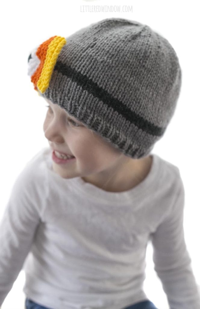 little girl in white shirt looking off to the left and wearing a gray knit hat with candy corn colored bow on the front
