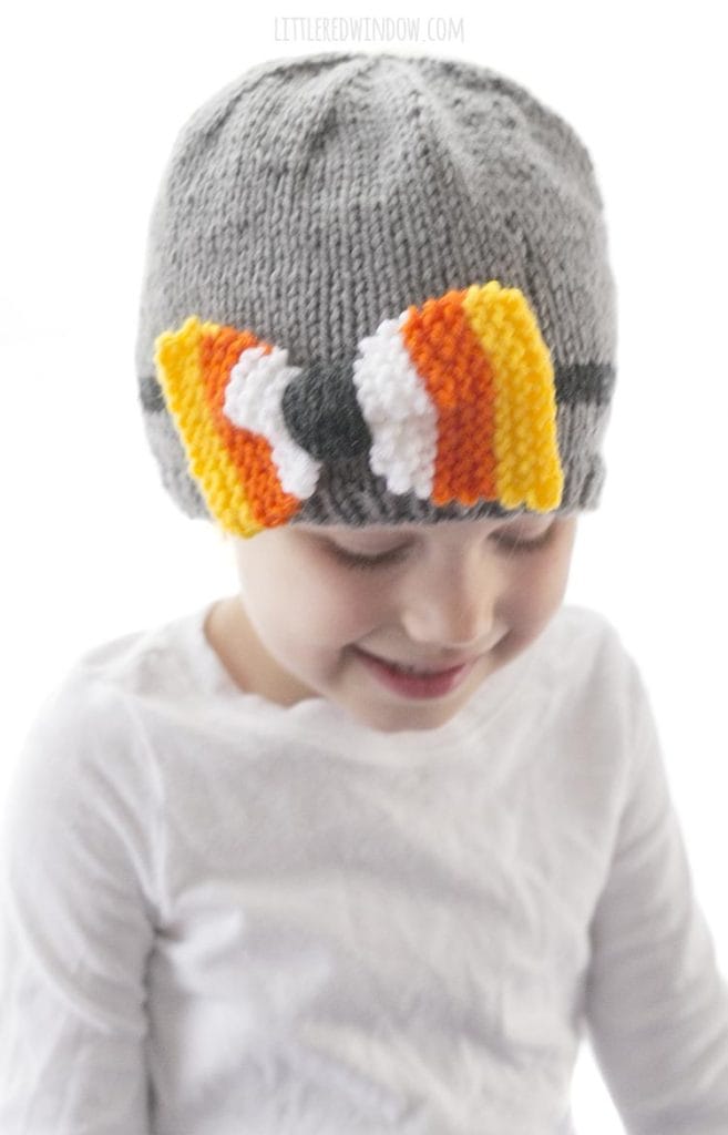 little girl looking down and wearing a gray knit hat with a bow striped yellow orange and white like candy corn
