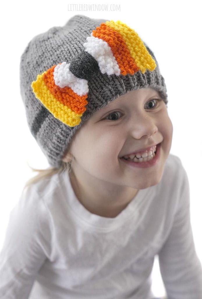 little girl in white shirt smiling and wearing a gray knit hat with candy corn colored bow on the front