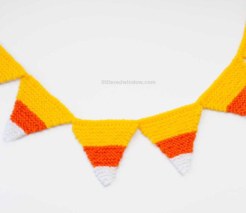 A candy corn garland of knitted triangles, each featuring white, orange, and yellow stripes, is displayed against a white background.