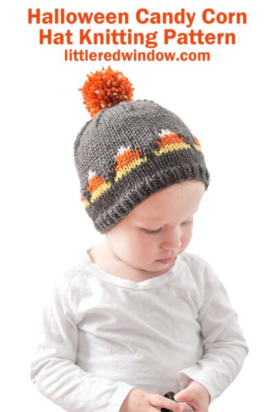 An infant wearing a grey knitted Candy Corn Hat featuring a candy corn pattern and a large orange pom-pom on top. The text above reads "Halloween Candy Corn Hat Knitting Pattern" and "littleredwindow.com." The infant is dressed in a white shirt and looking downward.