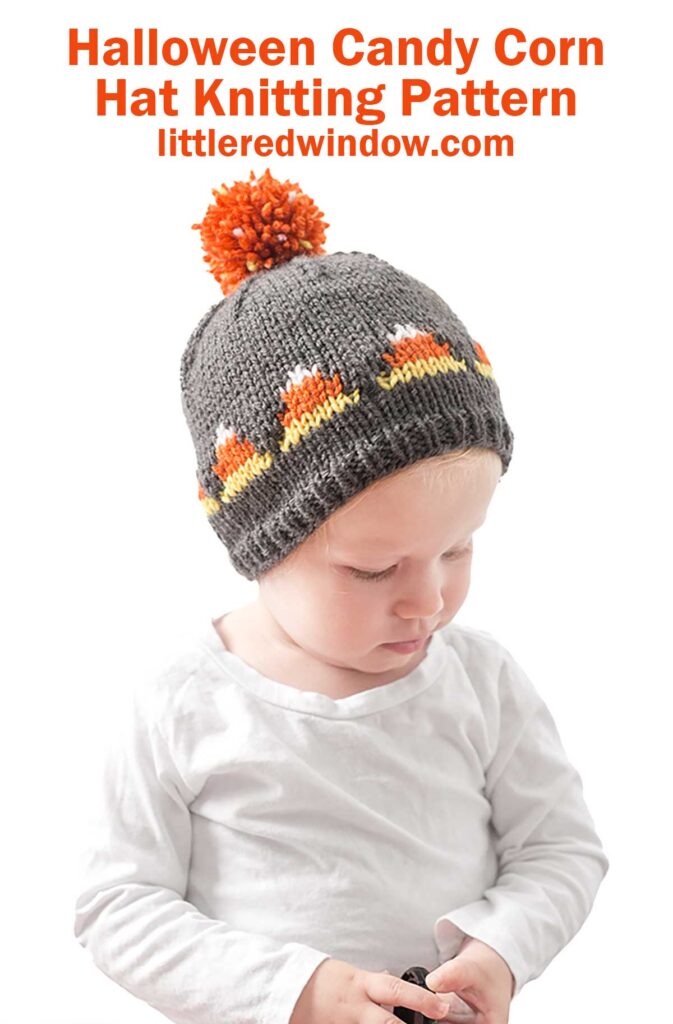 An infant wearing a grey knitted Candy Corn Hat featuring a candy corn pattern and a large orange pom-pom on top. The text above reads