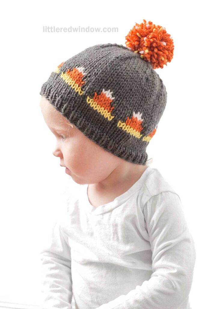 A toddler wearing a gray knitted candy corn hat with orange, white, and yellow accents and an orange pom-pom on top. The child is dressed in a white long-sleeve shirt and is looking downwards. The background is plain white.