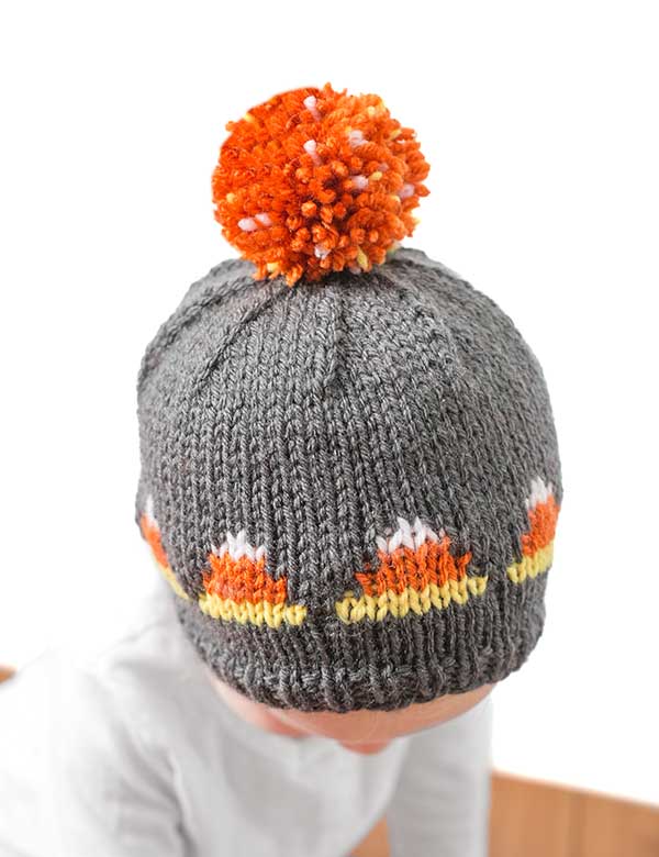 A baby wearing a knitted, dark gray candy corn hat decorated with an orange and white pattern resembling the festive treat. The top of the hat is adorned with a fluffy orange pom-pom. The background is plain white.