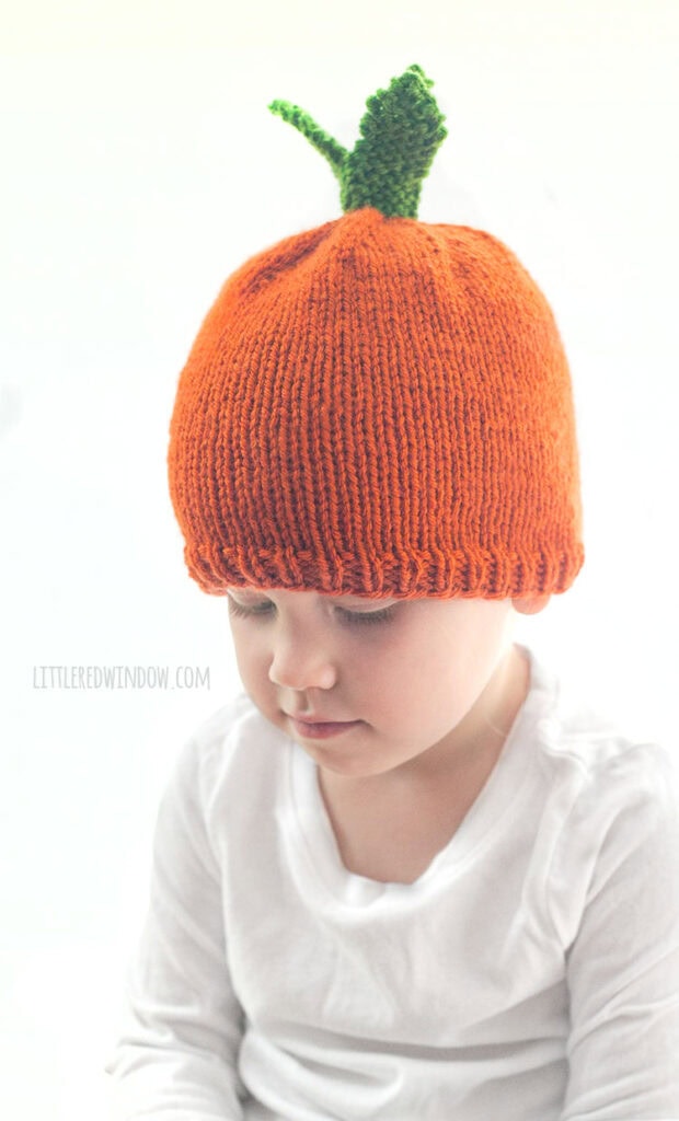 child in white shirt wearing an orange knit carrot hat with green knit carrot leaves on top looking to the left