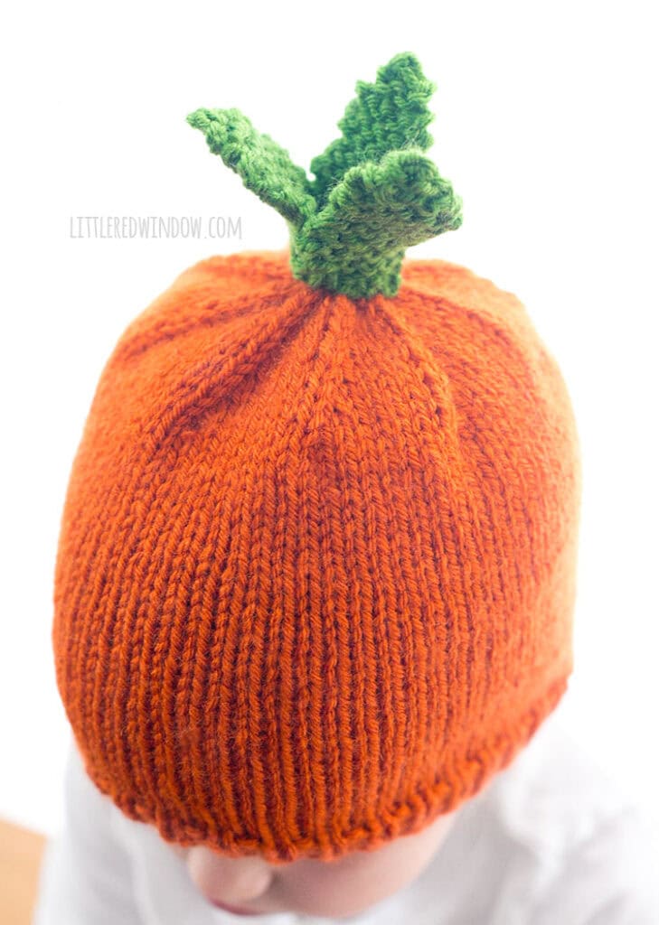 top view of child wearing a knit hat that looks like a carrot in front of a white background looking down to the left