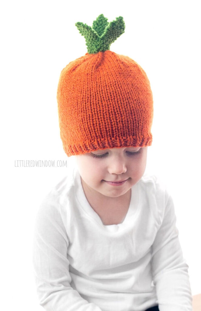 child in white shirt wearing an orange knit carrot hat with green knit carrot leaves on top looking down at their lap