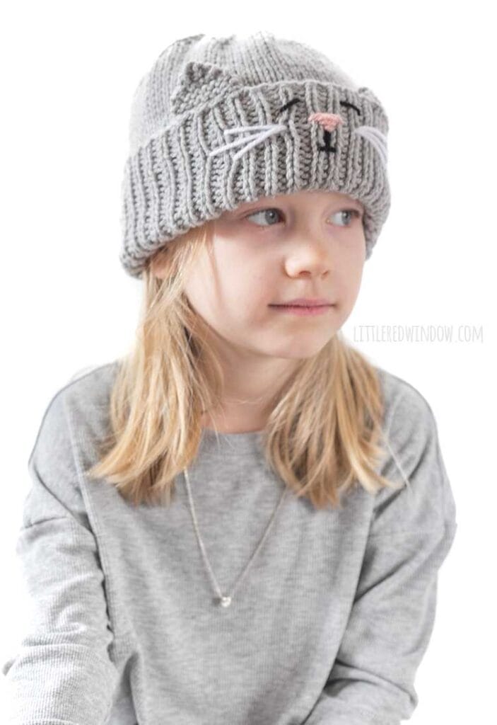 blond girl in gray shirt looking off to the right at her lap and wearing a gray knit hat with a cat face and ears embroidered into the thick ribbed folded brim