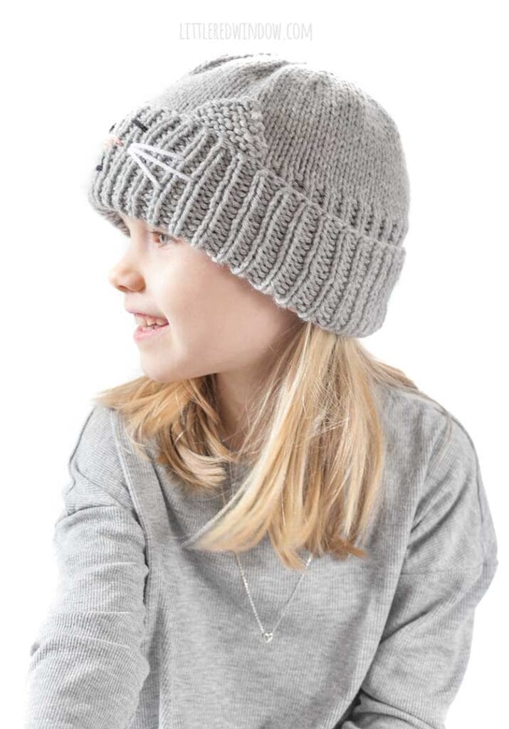 smiling blond girl in gray shirt looking off to the far left at her lap and wearing a gray knit hat with a cat face and ears embroidered into the thick ribbed folded brim