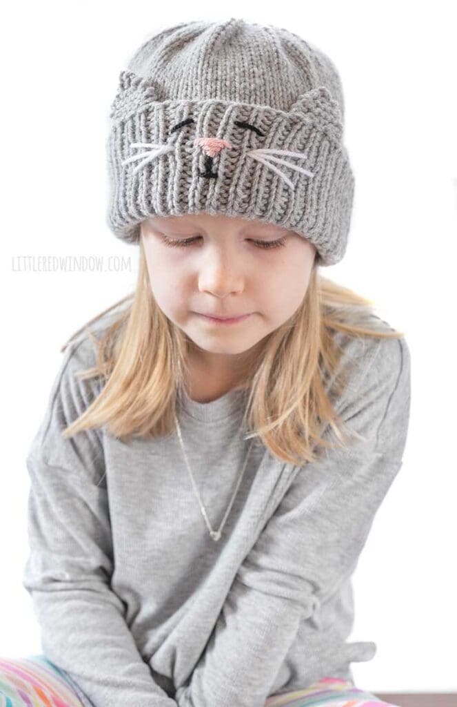 blond girl in gray shirt looking down at her lap and wearing a gray knit hat with a cat face and ears embroidered into the thick ribbed folded brim