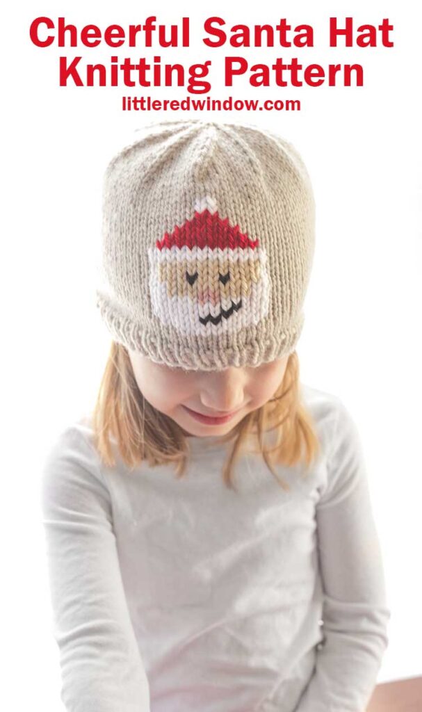 smiling girl looking down wearing a tan knit hat with a duplicate stitch Santa Face on the front of the hat with white beard smile and classic red Santa hat