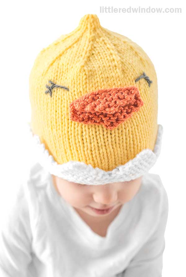 A child wearing a chick hat knitted in bright yellow, complete with embroidered eyes and an orange beak. The adorable hat boasts a white, fluffy trim. Dressed in a white outfit, the child stands against a plain background.