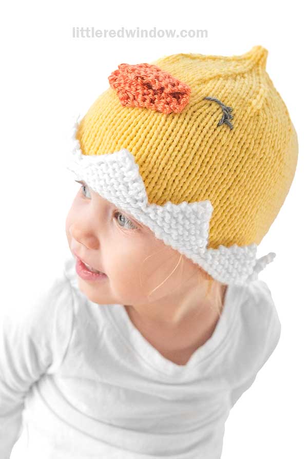 A young child wearing a chick hat, complete with a yellow knitted design and an orange beak, looks to the side in their white shirt. The whimsical egg shell detail adds to the cheerful expression on their face.
