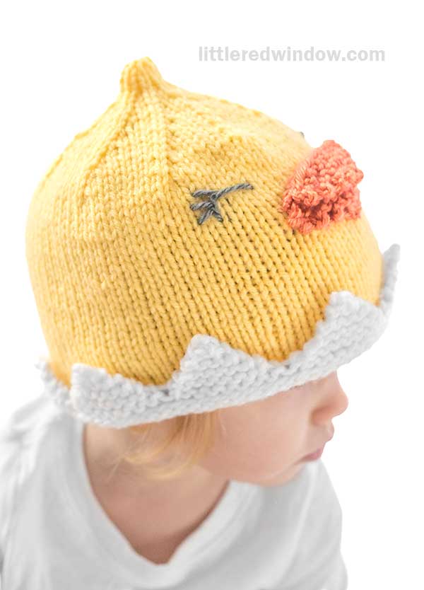 A child wearing a whimsical chick hat, featuring a knitted yellow design with an orange beak and gray eyes. The hat includes a playful white zigzag base resembling an eggshell, all set against a plain white background.