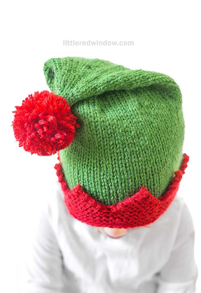 A child dons a charming knit green elf hat, crafted from an exquisite knitting pattern, complete with a red pom-pom and zigzag trim. Wearing a white shirt, they gaze downward in quiet contemplation.
