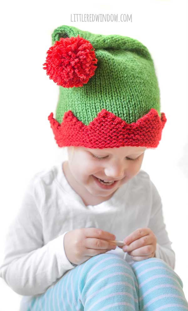 A child wearing a festive green and red elf hat, designed with a cozy knitting pattern, sits smiling in a white long-sleeve shirt and blue-striped pants against a crisp white background.