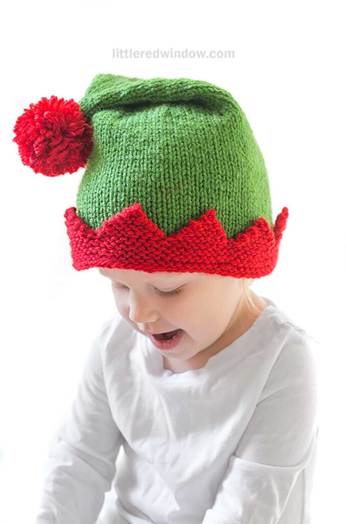 A child wearing a festive green elf hat, crafted from an intricate knitting pattern, features a red pom-pom and zigzag trim. Dressed in a white shirt against a plain white background, the child looks down, capturing the essence of holiday cheer.