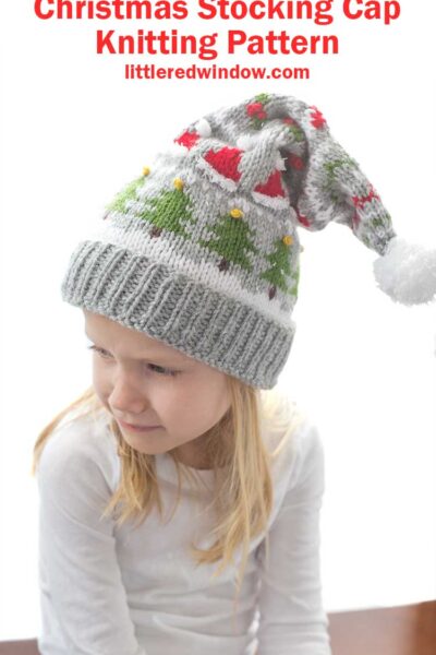 blond girl in white long sleeved shirt wearing a gray knit stocking cap decorated with patterns of Christmas trees Santa hats holly berries and candy cane stripe and a white pom pom on top looking down and off to her left