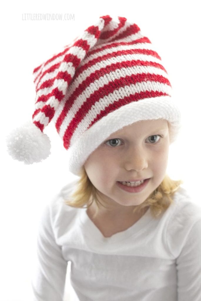 girl in white shirt lwearing a red and white striped knit stocking cap with white rolled brim and white pom pom looking off to the right