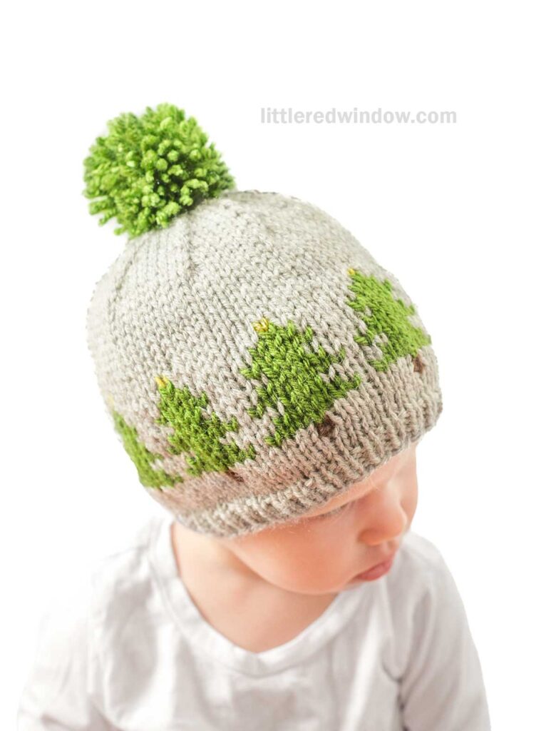 A baby wears a cozy gray knitted hat adorned with a green Christmas tree design around the brim and topped with a playful pom-pom. The little one gazes downward, set against a plain white backdrop, evoking the charm of a serene Christmas Tree Farm scene.