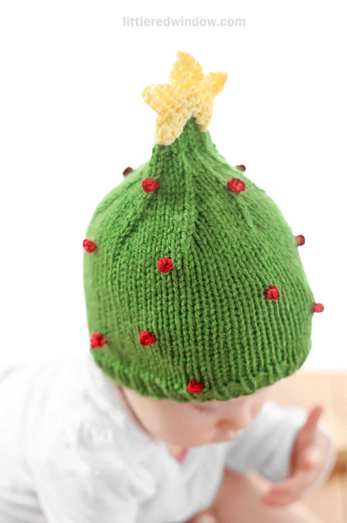 A baby in a Christmas tree hat, knitted green with small red dots and a yellow star on top, is dressed in white. The adorable scene captures the holiday spirit as the baby, partially visible and facing downward, charms everyone with festive cuteness.
