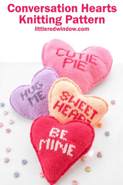 Four knitted hearts in pink, orange, red, and purple, each with a playful word or phrase like "CUTE PIE" and "BE MINE" stitched on them. They're surrounded by small, colorful candy conversation hearts. Text above reads, "Conversation Hearts Knitting Pattern.