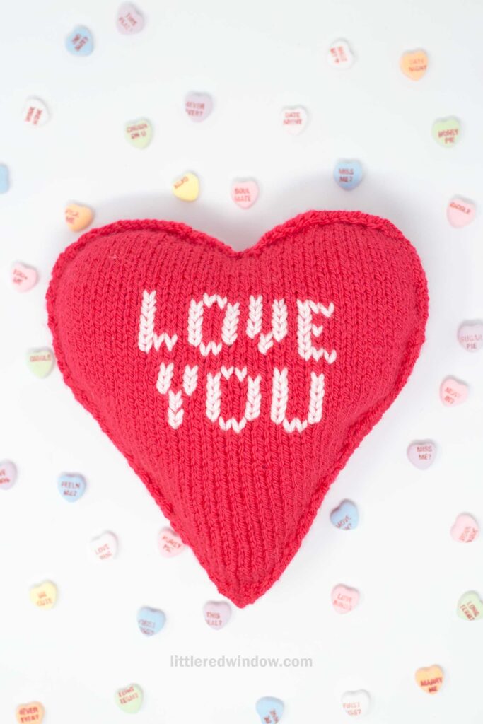 A red knitted heart-shaped pillow with "LOVE YOU" stitched in white text. It is surrounded by small, colorful heart-shaped candies with various messages on them. The background is a light, neutral color.