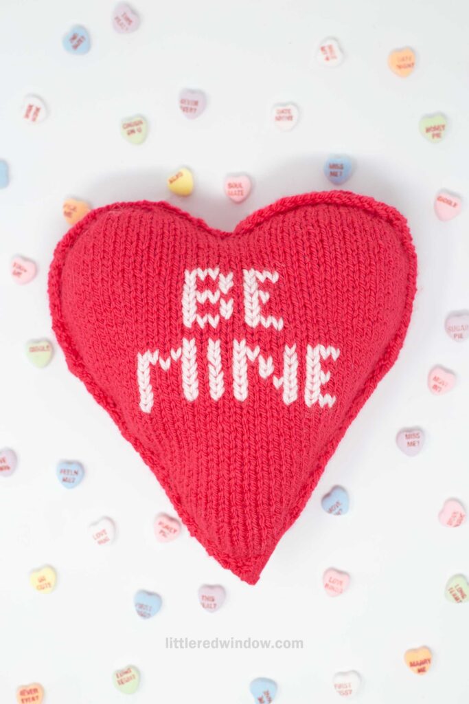A red knitted heart-shaped pillow with "BE MINE" stitched in white text. It is surrounded by small, colorful heart-shaped candies with various messages on them. The background is a light, neutral color.