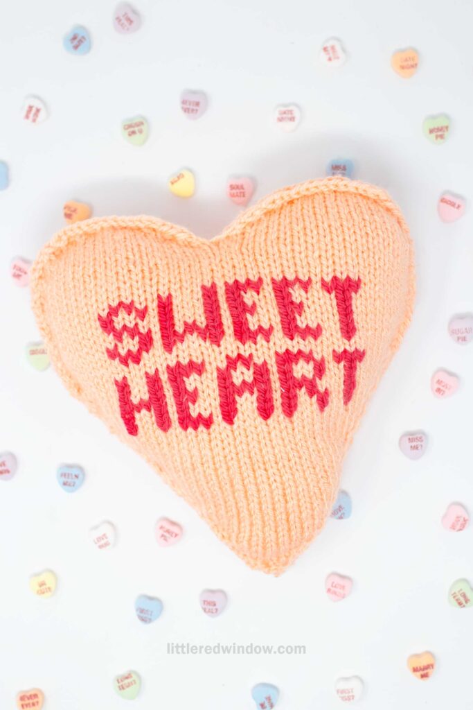A knitted heart-shaped pillow with "Sweet Heart" in red letters on a soft orange background, surrounded by scattered pastel-colored candy hearts.