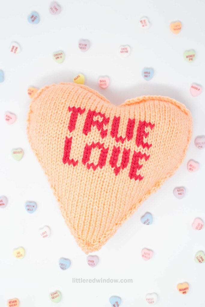 A light orange knitted heart-shaped cushion with "TRUE LOVE" stitched in red. It is surrounded by pastel-colored candy hearts with various words on them. The background is a plain white surface.