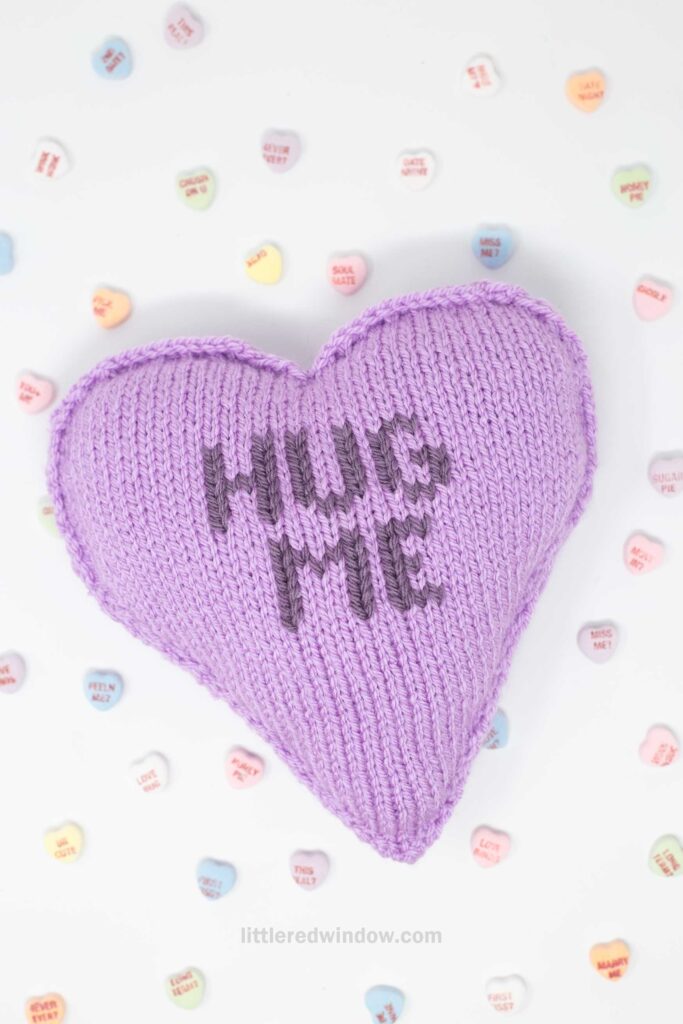A knitted purple heart pillow with "HUG ME" in dark purple letters, surrounded by pastel-colored candy hearts scattered on a white background.