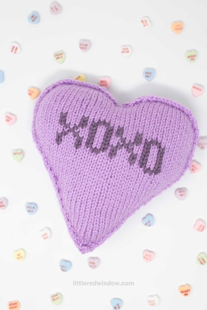 A knitted purple heart pillow with "XOXO" stitched on it is surrounded by small, colorful conversation candy hearts. The background is white, creating a light and cheerful scene.