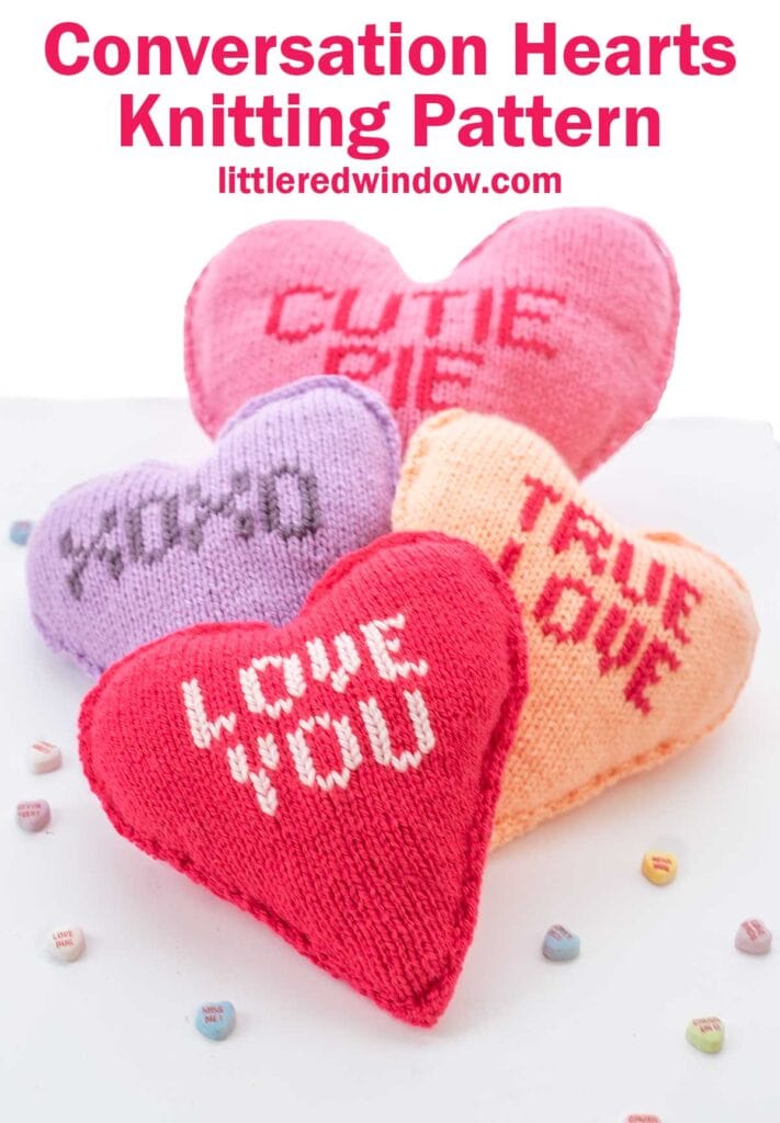 Four knitted heart-shaped cushions in pink, purple, red, and peach are displayed with phrases like "CUTIE PIE" and "LOVE YOU." Tiny candy hearts are scattered around them. The text reads "Conversation Hearts Knitting Pattern littleredwindow.com.