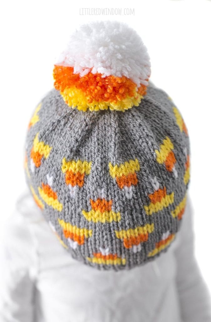 Closeup top view of round candy corn colored yarn pom pom on top of a gray knit baby hat with a fair isle candy corn pattern all over it