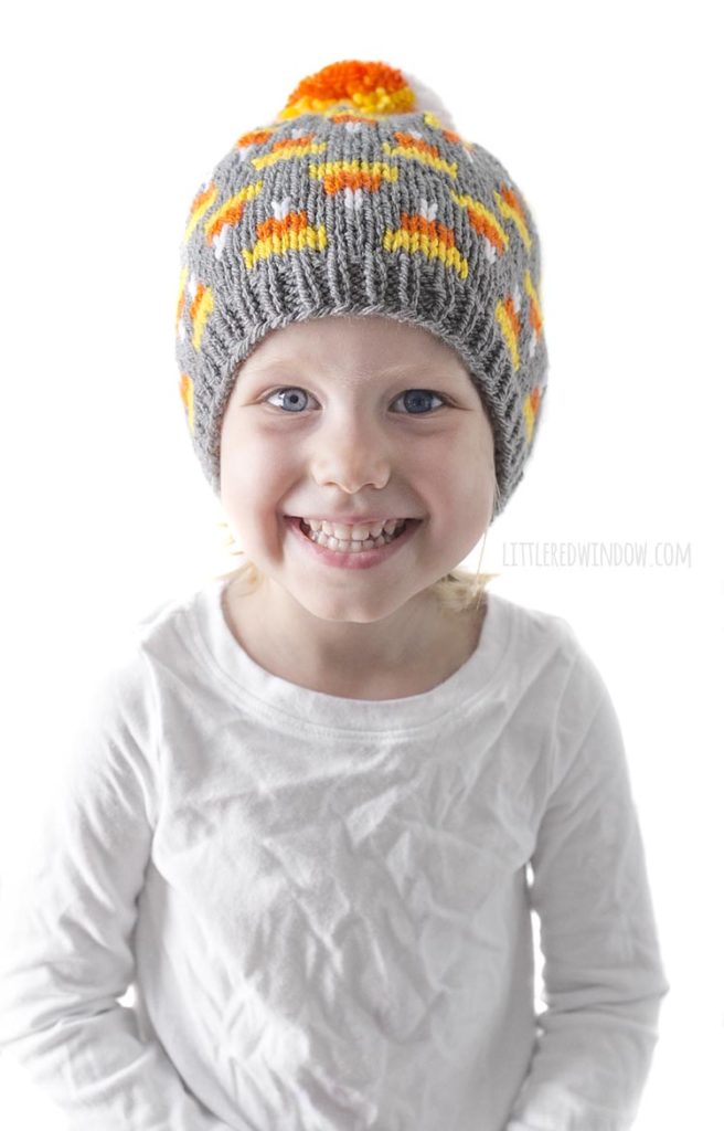 little girl in white shirt smiling and wearing gray knit hat with candy corn pattern on it and candy corn colored pom pom on top