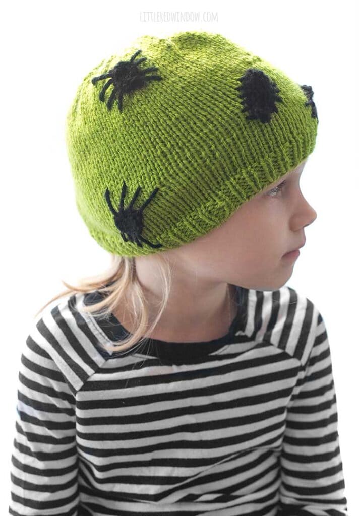 child in black and white striped shirt wearing an acid green knit hat with black knit spiders and bugs on it and looking off to the right in front of a white background