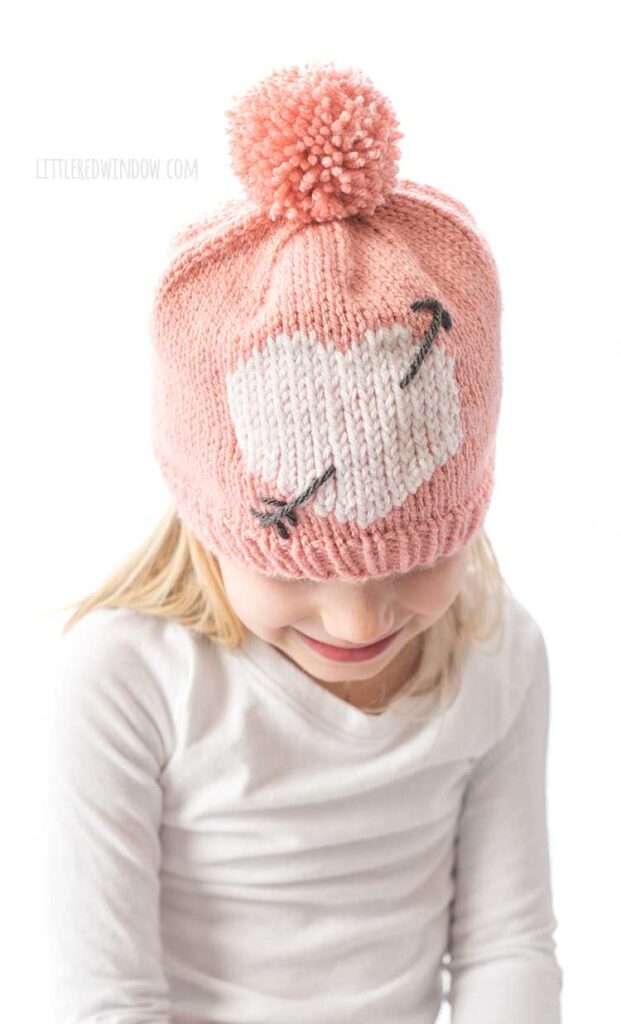 girl in white shirt looking down and wearing a light pink knit hat with large pink pom pom on top and a white heart with a charcoal gray arrow through it on the front of the hat