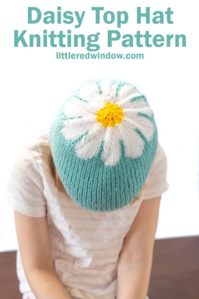 child leaning forward to show the top of a blue knit hat with a white and yellow daisy pattern centered on the top of it