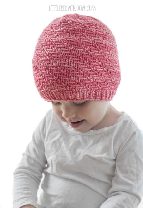 toddler in white shirt wearing coral pink knit hat with diagonal weave stitch pattern looking down
