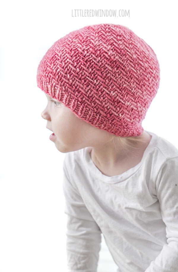 girl in white shirt wearing bubblegum pink knit hat with diagonal weave knit pattern on it looking off to the left