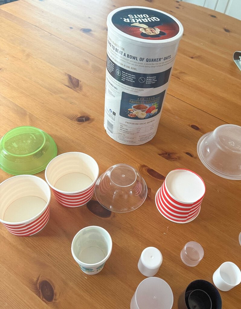 A tabletop with stacked cups and bowls, some in red and white stripes, others transparent, alongside a large oats container. Various small lids and a green bowl lid are scattered around.